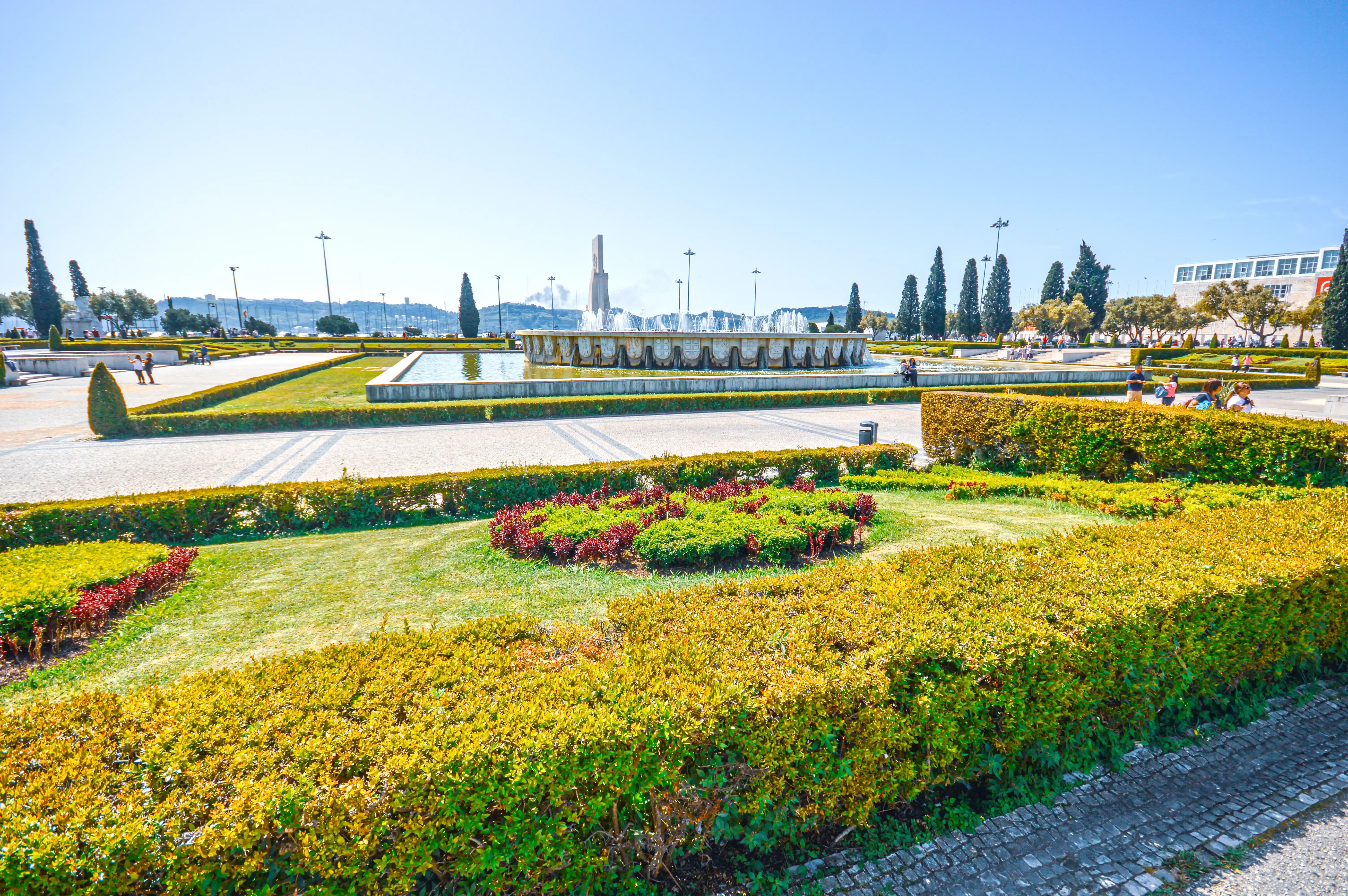 A wide shot of a large, circular fountain in full spray, surrounded by meticulously kept hedges and flowerbeds in a public park. Several people are visible walking and standing in the park on a sunny day with a clear blue sky.